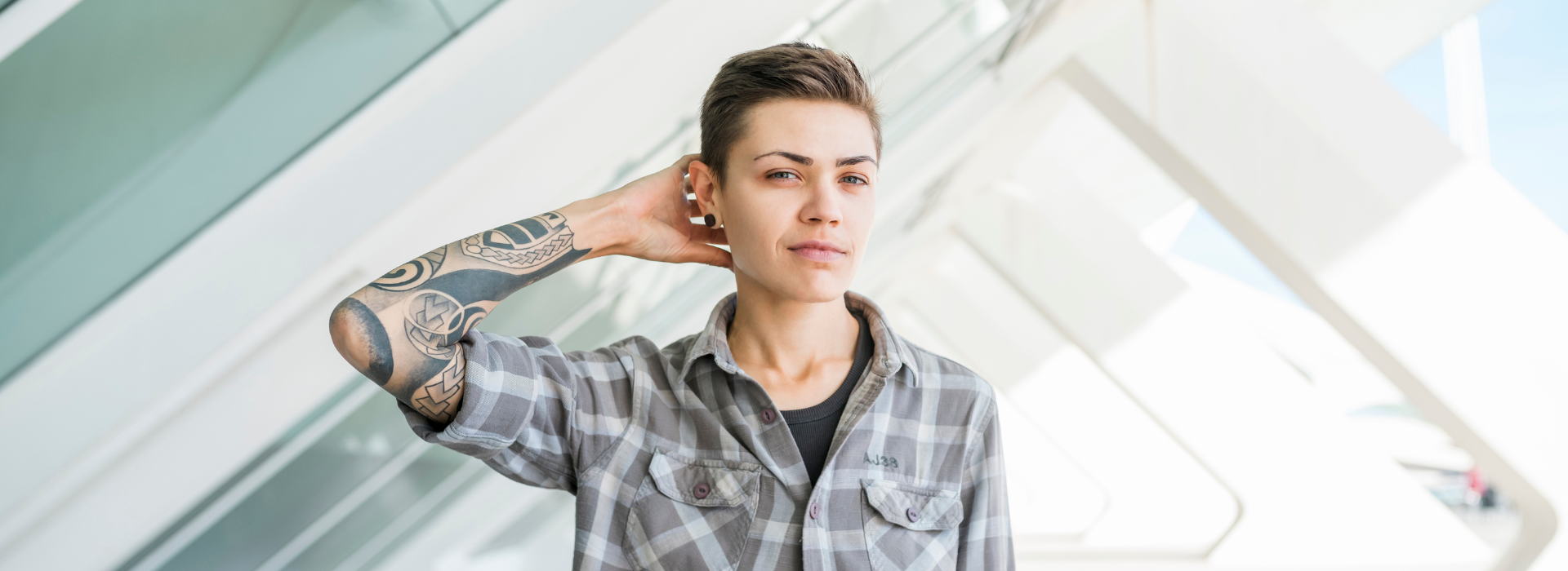 A transgender student with tattoos on their right arm and a plaid button down shirt glance at the camera.